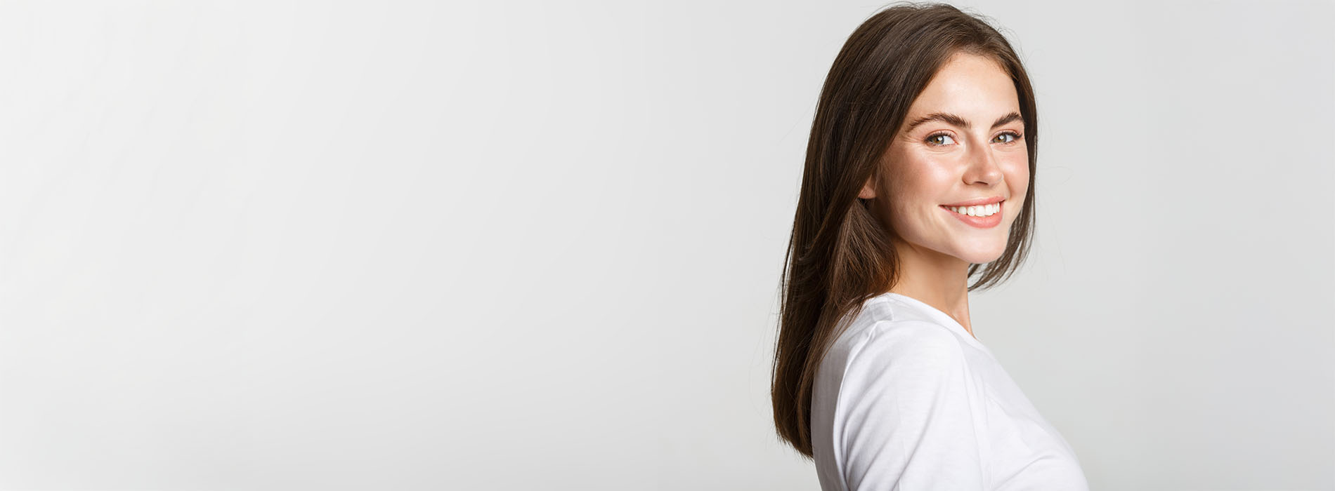 The image shows a woman standing against a white background, looking off to the side with a slight smile, wearing a light-colored top.